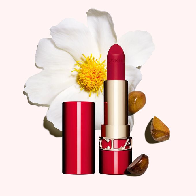 Plan produit du Joli Rouge Velvet de Clarins en teinte 742V, à côté d’un camélia et de graines de karité sur fond rose.
